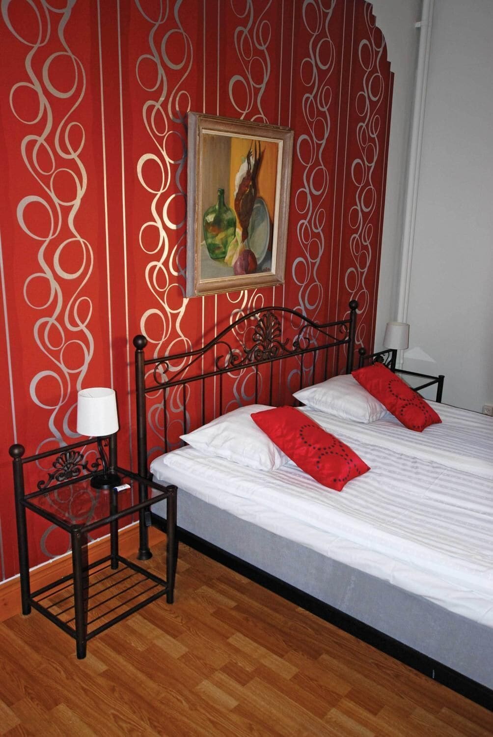 Una cama bien hecha con almohadas blancas y rojas, un cuadro en la pared y una mesita de noche con una lámpara.