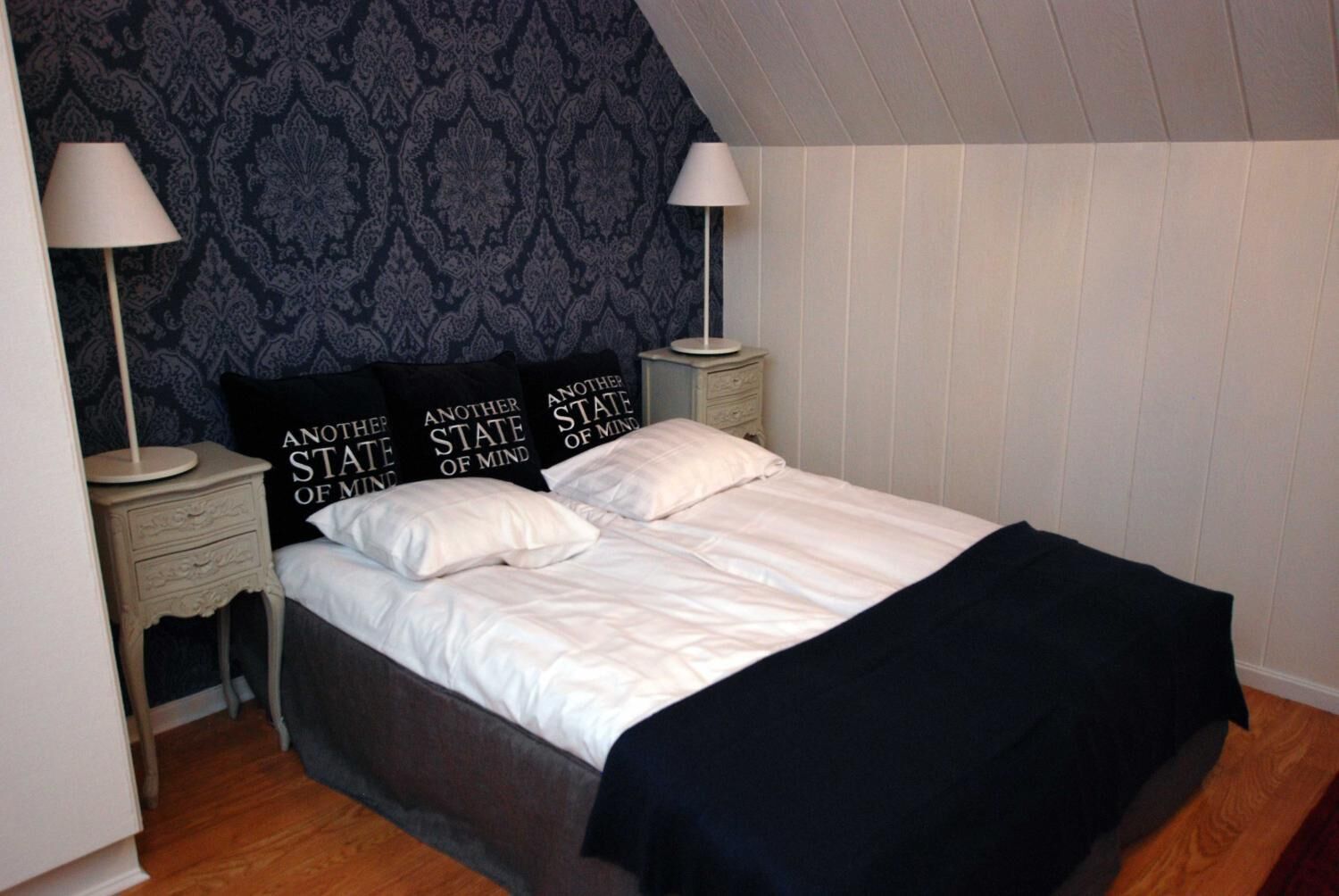 A neatly made bed with white linens and a black throw blanket, flanked by two bedside tables with lamps, in a room with patterned wallpaper and wooden paneling.