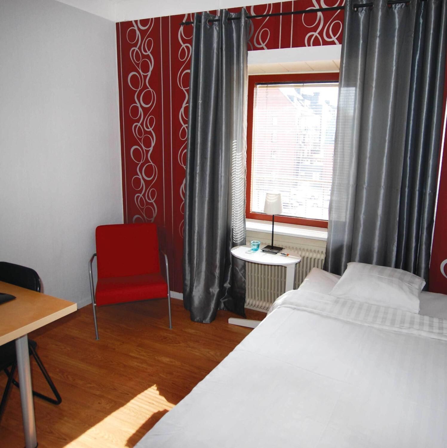 Une chambre d’hôtel avec un lit, une chaise rouge, une petite table avec une lampe et une fenêtre avec des rideaux.