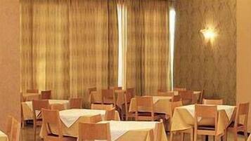 A hotel dining area with multiple tables and chairs, large windows with curtains, and a dark-colored door.