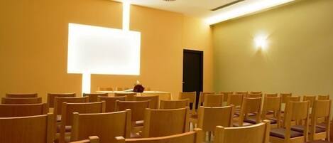 A conference room with rows of wooden chairs and a large white screen at the front.