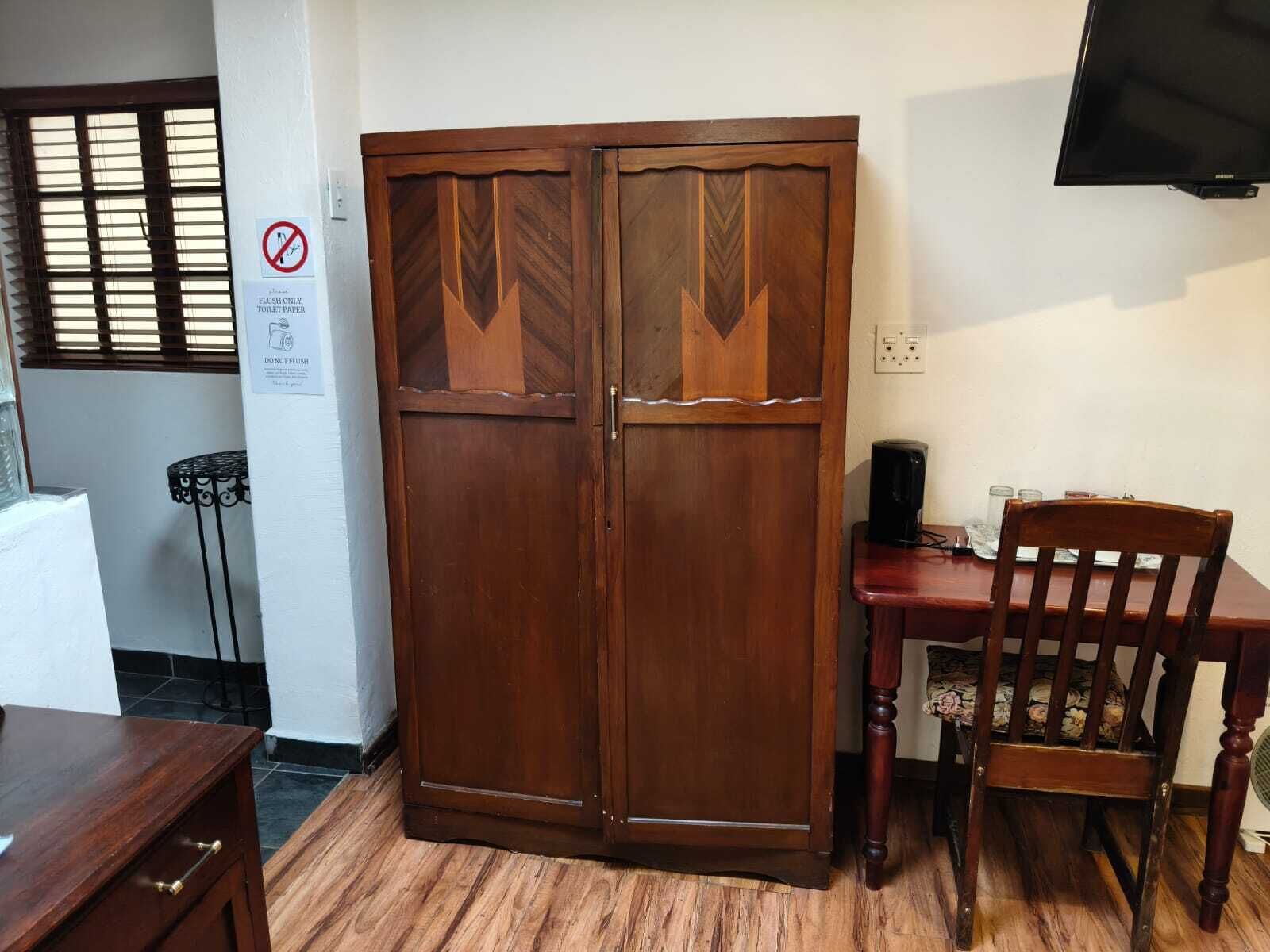 A room with a wooden wardrobe, a desk with a chair, a television mounted on the wall, and a sign indicating the use of toilet paper.