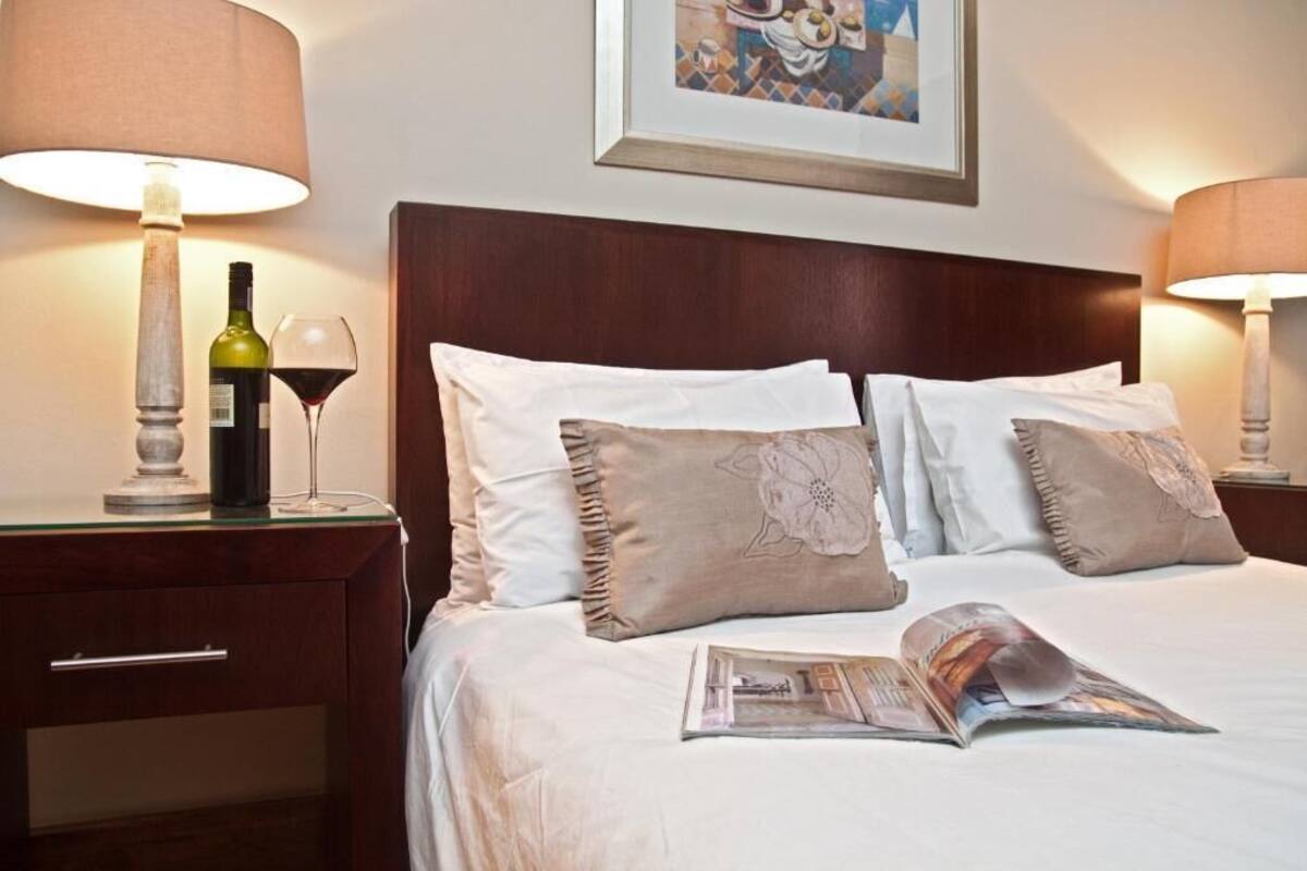 A neatly made bed with a magazine, a bottle of wine, and a glass on a bedside table.