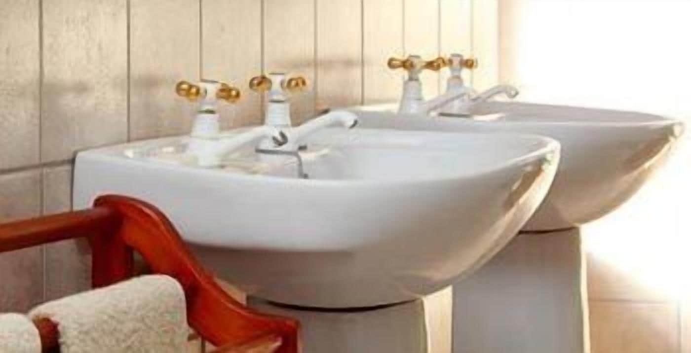 Dos lavamanos blancos sobre pedestal con grifos dorados en un baño con paneles de madera.