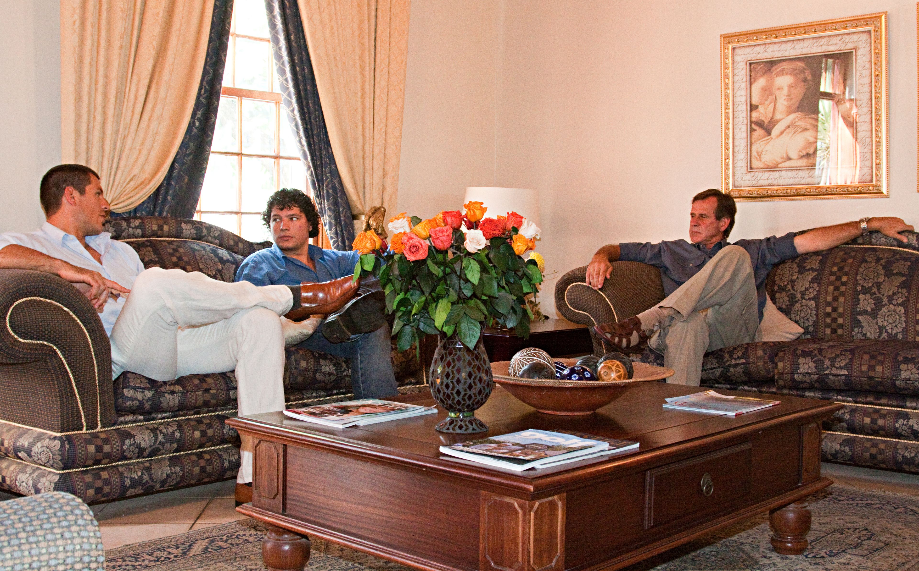 A living room with three people seated on patterned sofas, a wooden coffee table with magazines, a vase with flowers, and a framed picture on the wall.