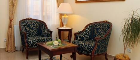 Une pièce comprenant deux fauteuils richement décorés, une petite table sur laquelle est posé un vase de fleurs, une lampe et une plante en pot.