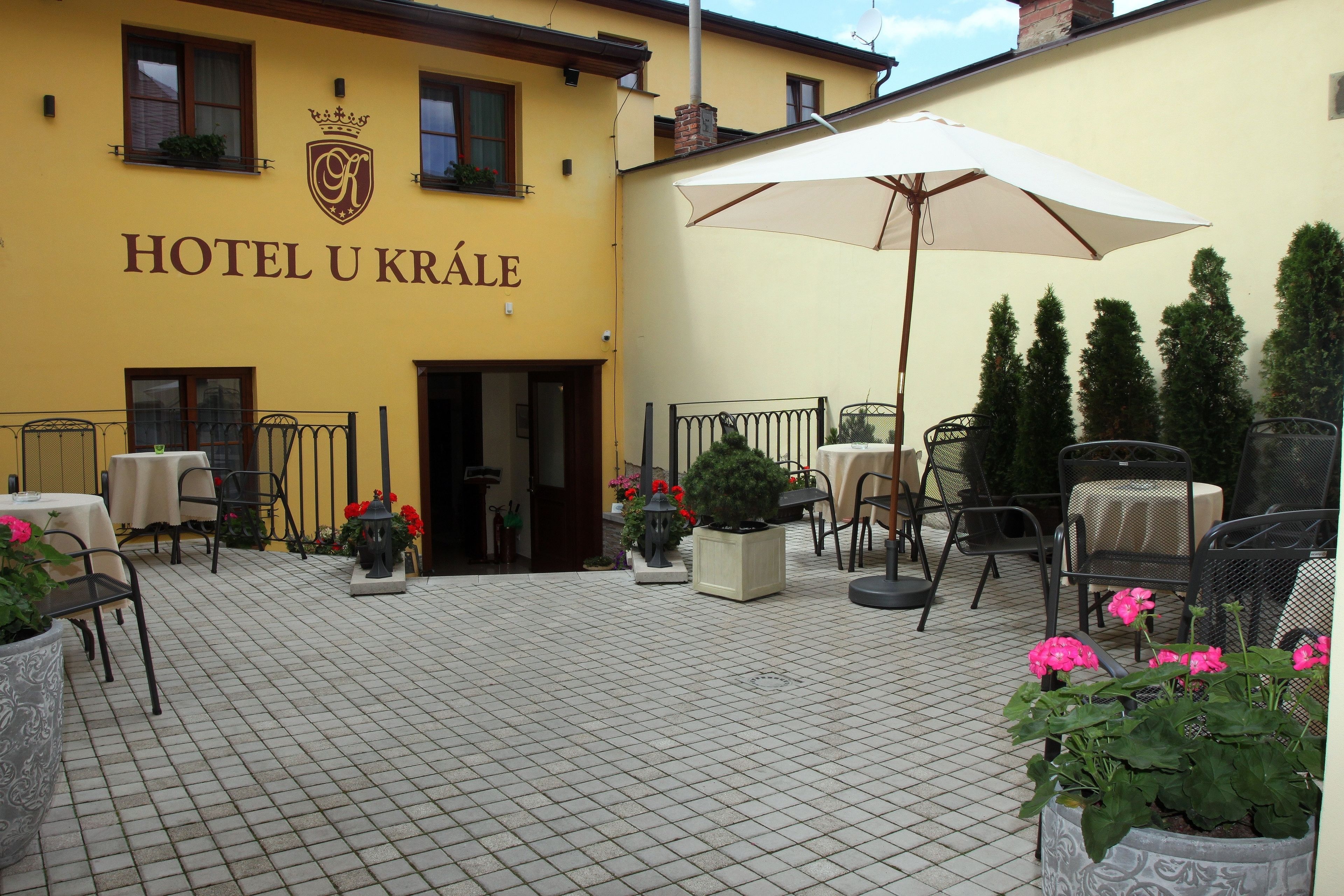A courtyard with a yellow building labeled "HOTEL U KRALE," featuring outdoor seating, potted plants, and a large umbrella.