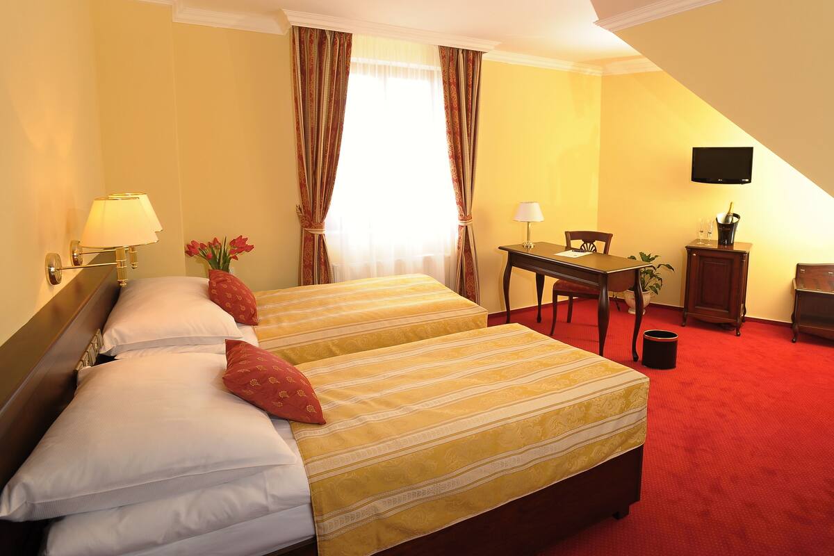 A hotel room with a double bed, two bedside tables with lamps, a desk with a chair, a television mounted on the wall, and a red carpeted floor.