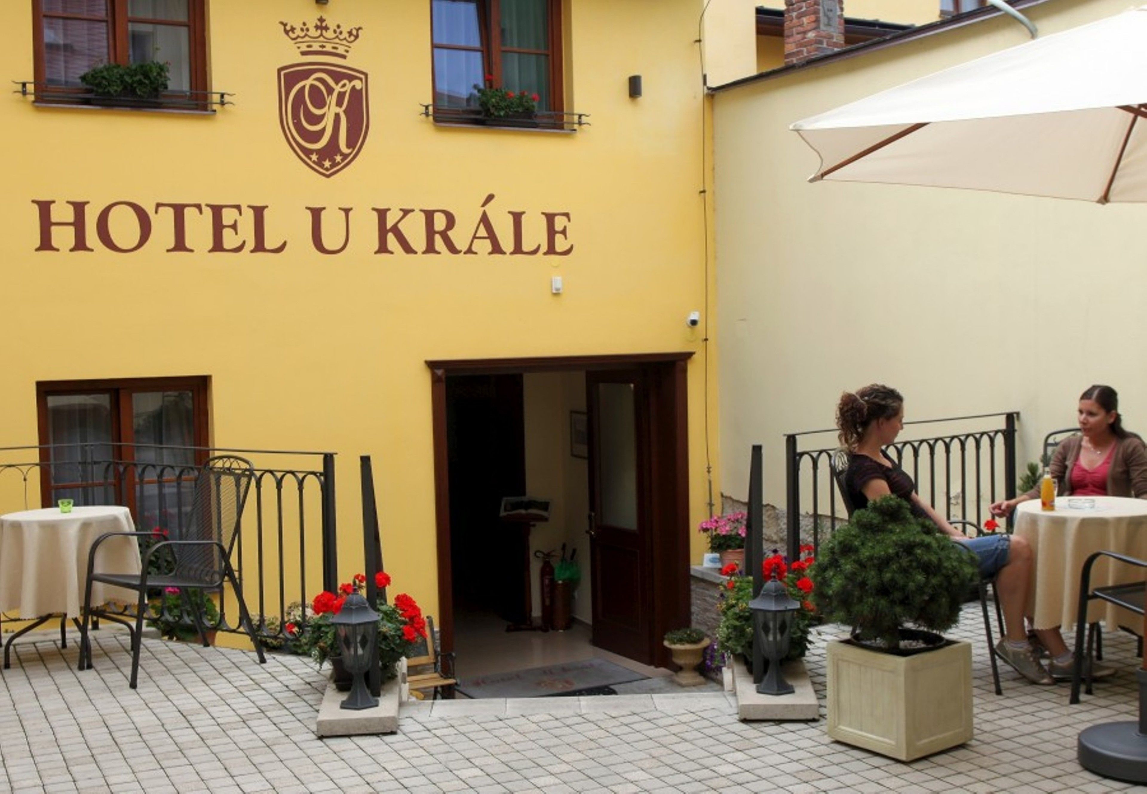 A yellow building with the sign "HOTEL U KRÁLE" and a crown emblem. There are outdoor tables and chairs, potted plants, and a large umbrella.
