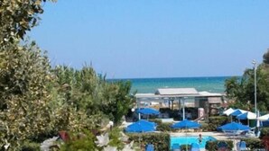 A table with a bottle, wine glasses, and a bowl of salad, overlooking a pool and beach area. - Galeana Beach Hotel (Rethymno)