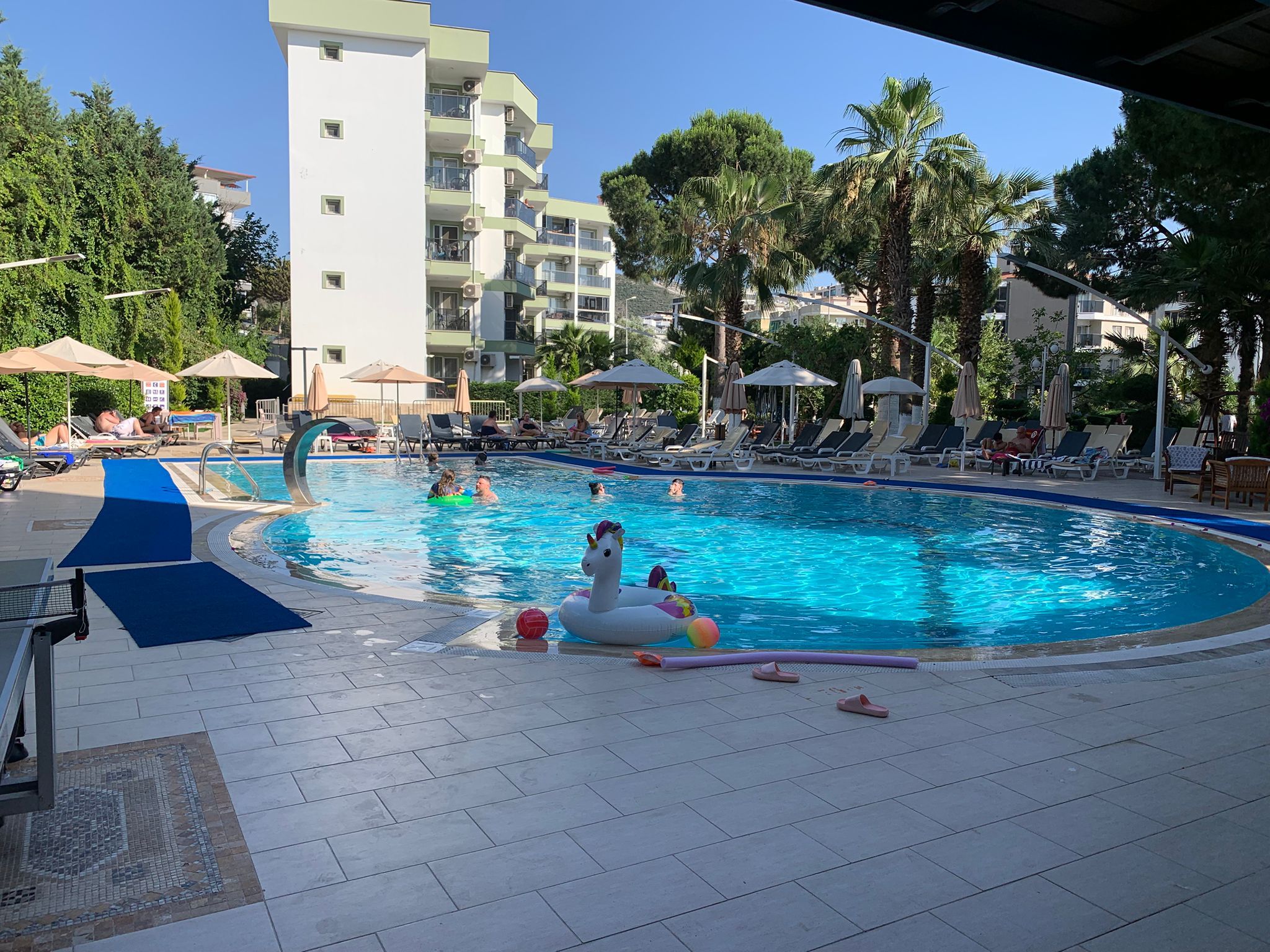Ein Schwimmbecken mit Liegestühlen und Sonnenschirmen, umgeben von Palmen und einem mehrstöckigen Gebäude.