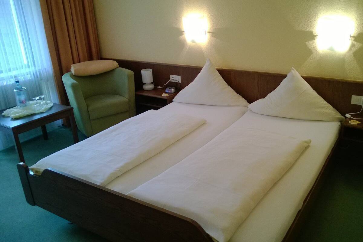 Ein Hotelzimmer mit einem Doppelbett, zwei Nachttischen, einem Sessel und einem Beistelltischchen mit Lampe.