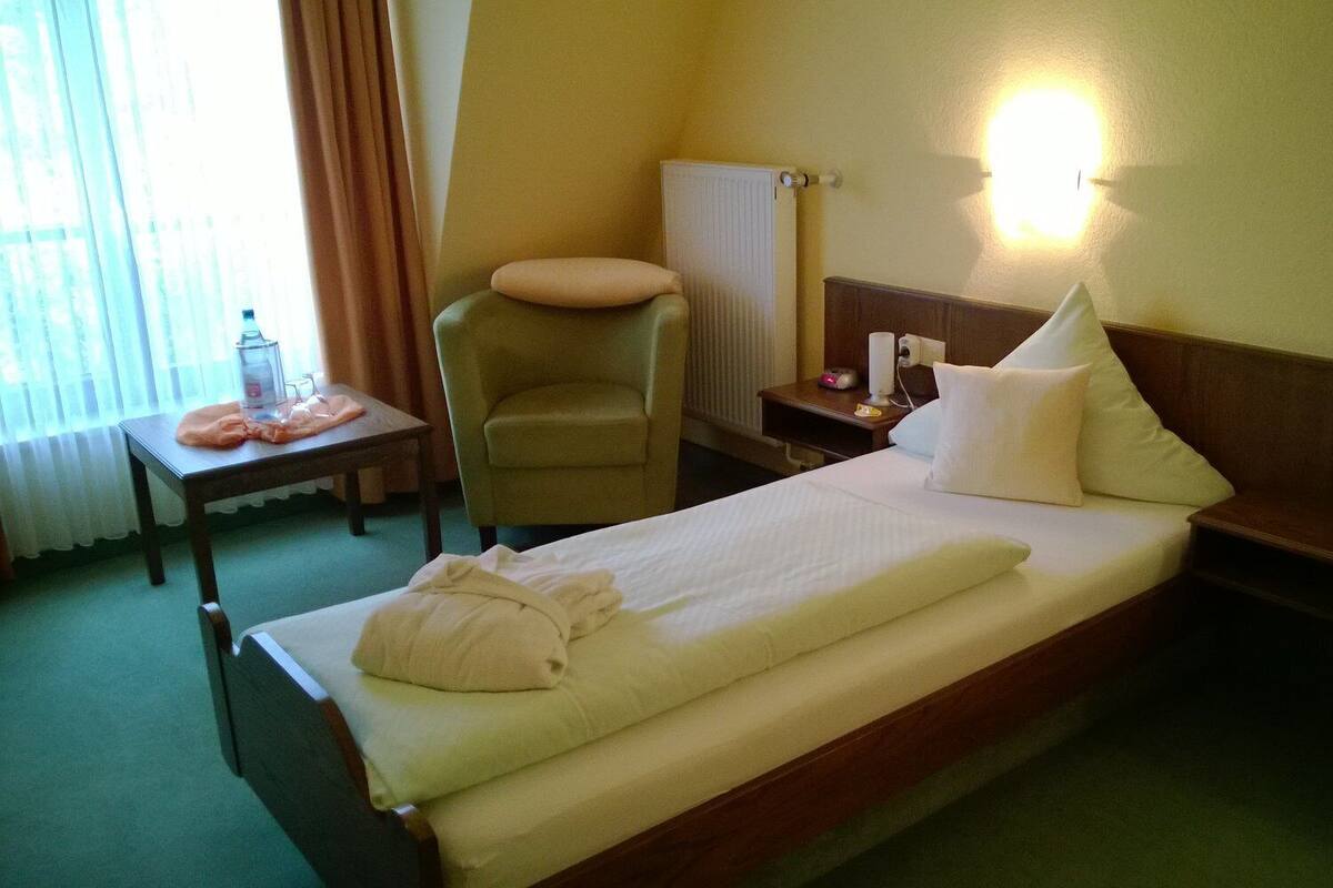 Ein Hotelzimmer mit einem Bett, einem Sessel, einem Tisch mit einer Flasche sowie einem Nachttisch mit einer Lampe und einem Blumenarrangement.