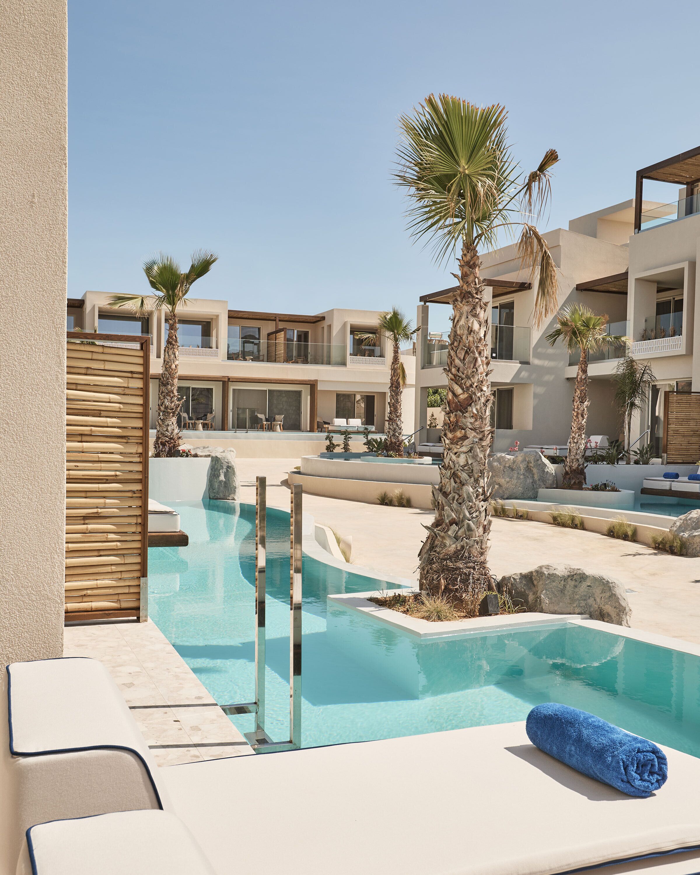 Un espace piscine avec des chaises longues et une serviette bleue, entouré de palmiers et de bâtiments modernes.