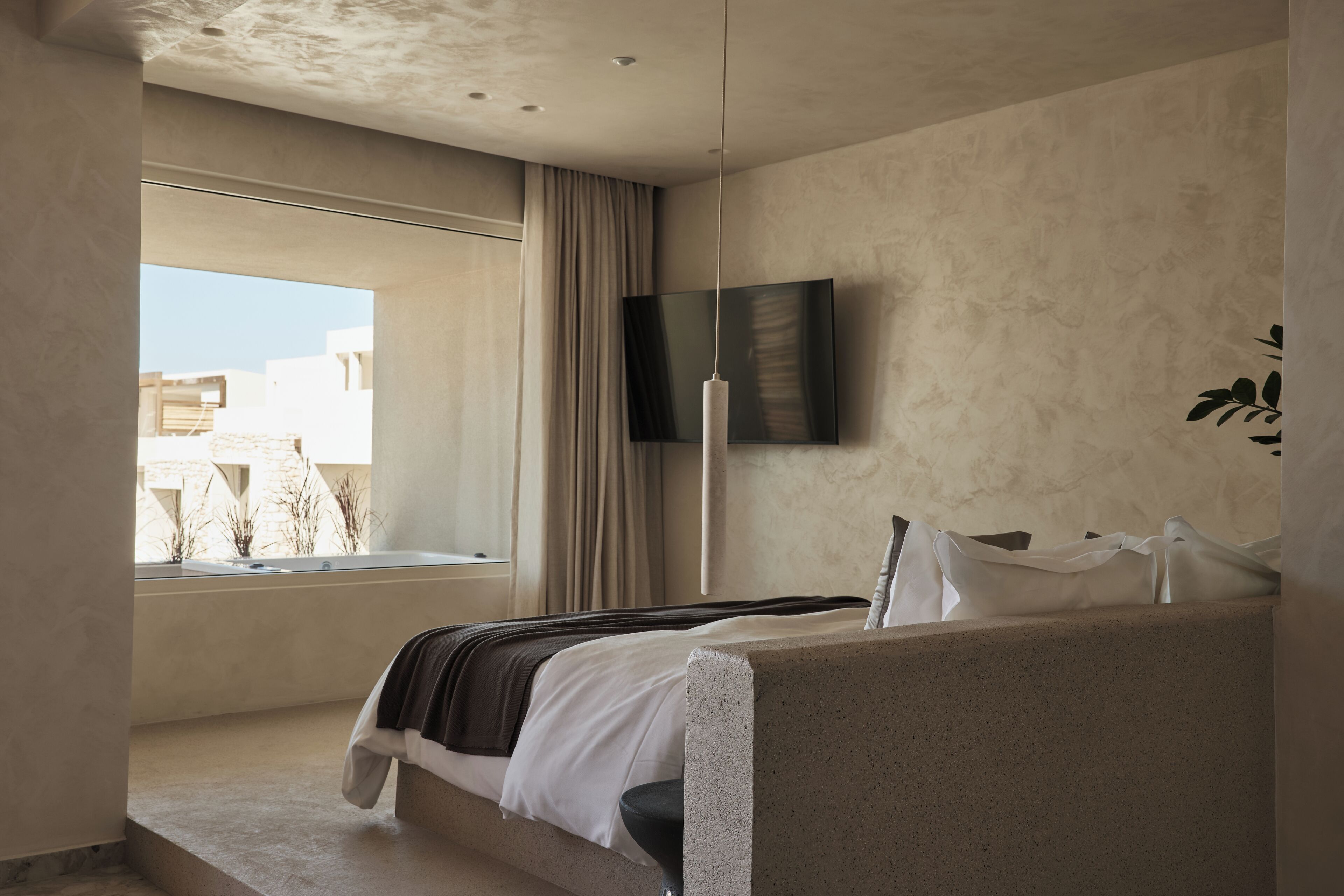 A modern bedroom with a large window, a flat-screen TV mounted on the wall, and a bed with a headboard.