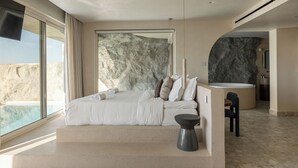 Un dormitorio con una cama grande, vista al océano, una pared de piedra y una mesita con una silla.