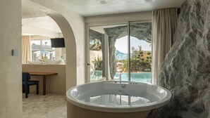 A modern bathroom with a large bathtub, a window with a view of a pool and palm trees, and a stone accent wall. - Uzenie All Suites Boutique Resort - Adults Only (Rethymno)