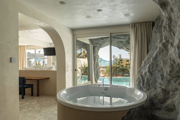 A modern bathroom with a large bathtub, a window with a view of a pool and palm trees, and a stone accent wall. - Uzenie All Suites Boutique Resort - Adults Only (Rethymno)