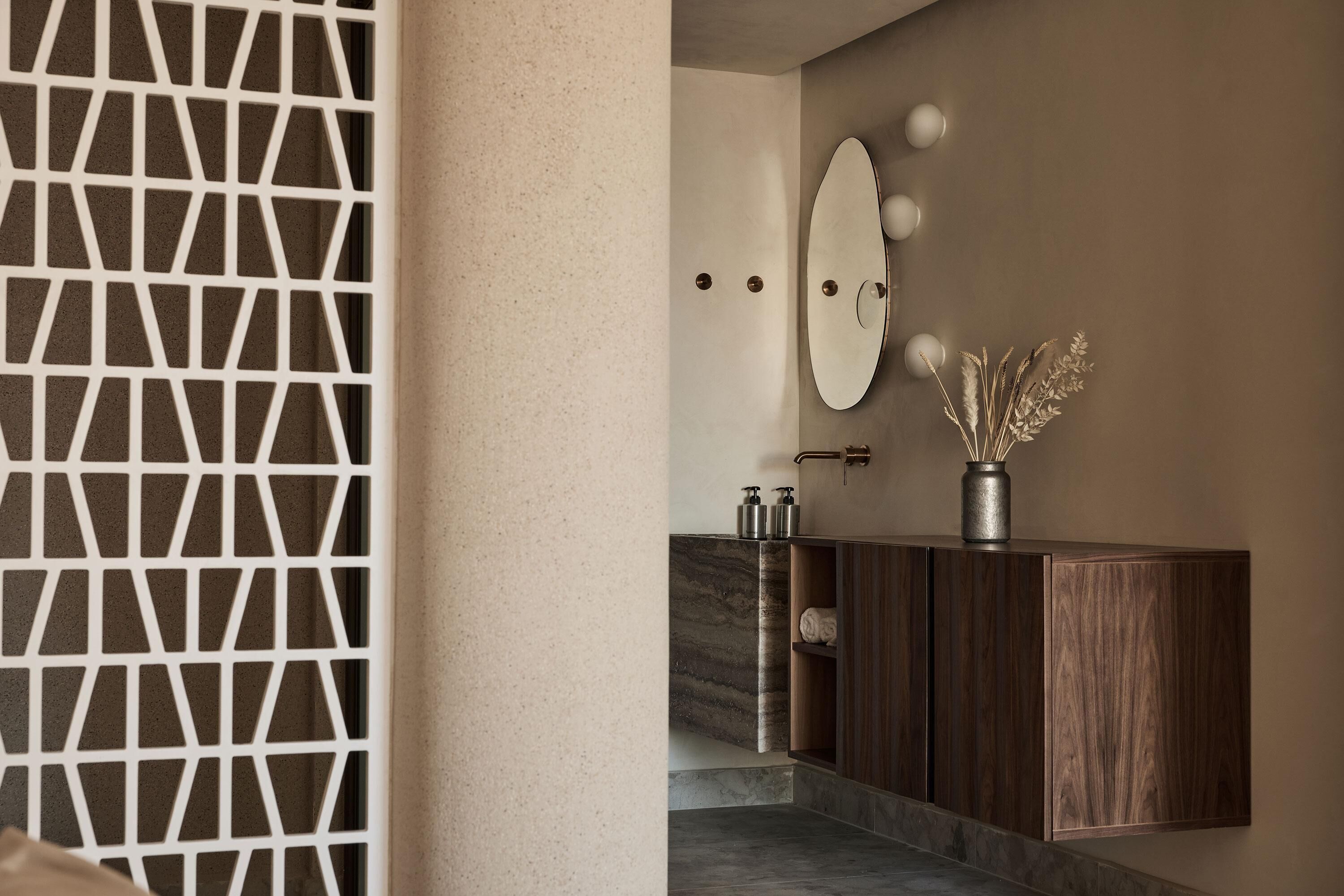Un baño moderno con una puerta de diseño geométrico, encimera de mármol, tocador de madera, espejo con iluminación y un jarrón decorativo con plantas secas.