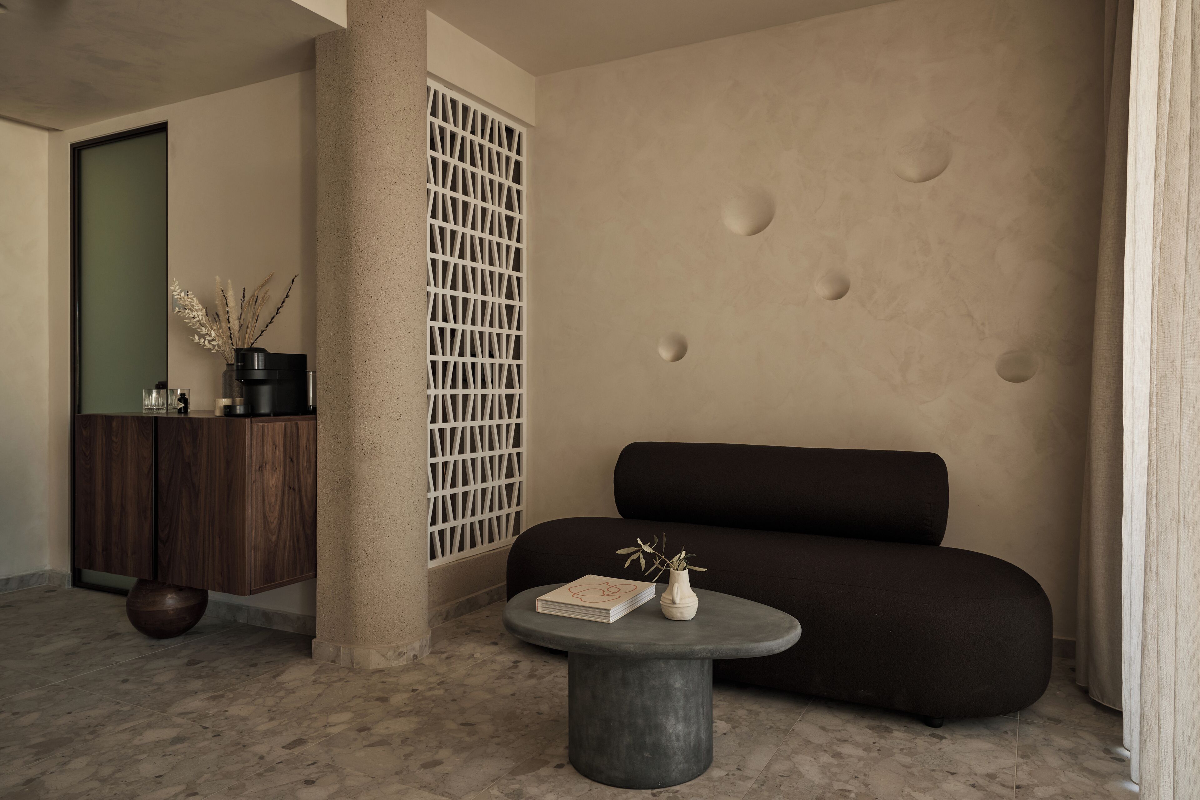 Ein modernes Interieur mit einem dunklen Sofa, einem runden Tisch mit Buch und Vase sowie einem Holzschrank.