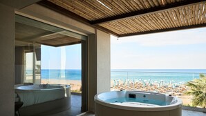 A room with a large window overlooking a beach, a jacuzzi, and a separate bathtub. - Uzenie All Suites Boutique Resort - Adults Only (Rethymno)