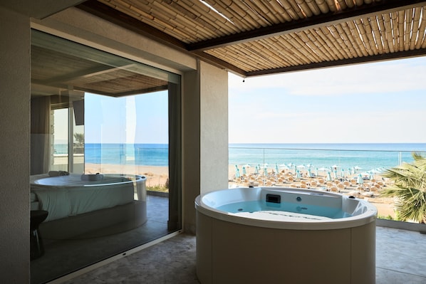 A room with a large window overlooking a beach, a jacuzzi, and a bed. - Uzenie All Suites Boutique Resort - Adults Only (Rethymno)