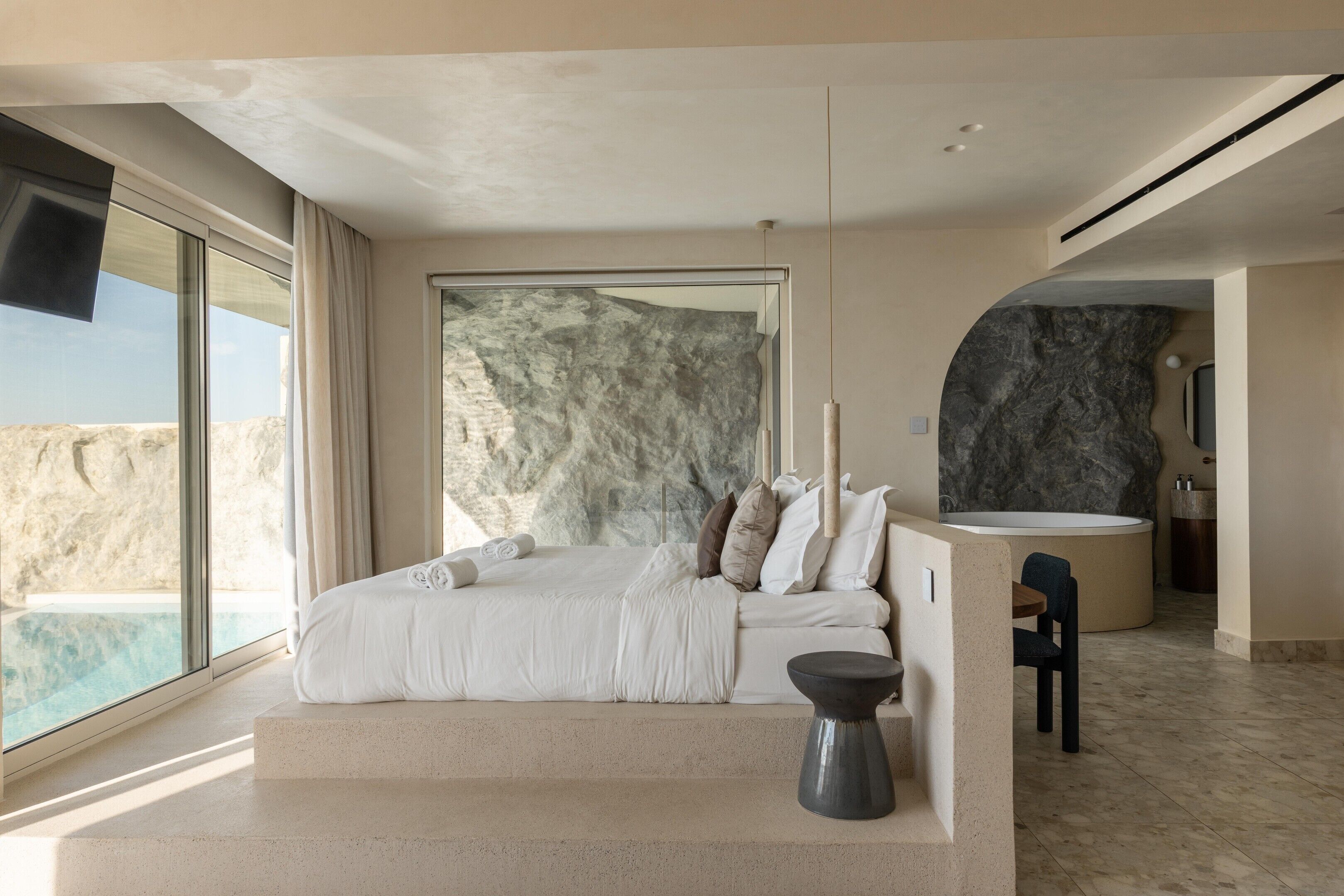 A modern bedroom with a large bed, a stone wall, and a view of the ocean.