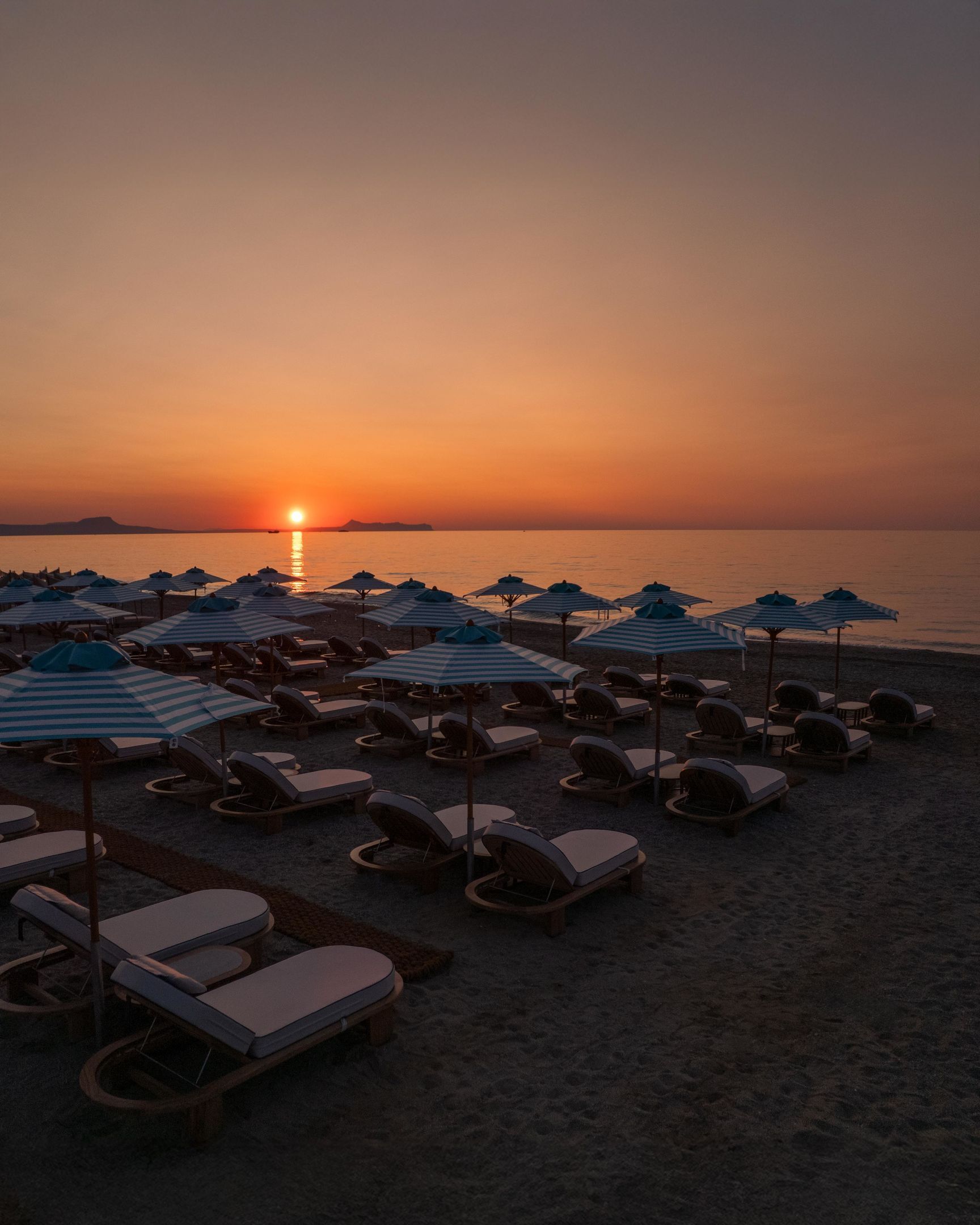 Una playa con filas de tumbonas y sombrillas a rayas, un atardecer de fondo y mar en calma.