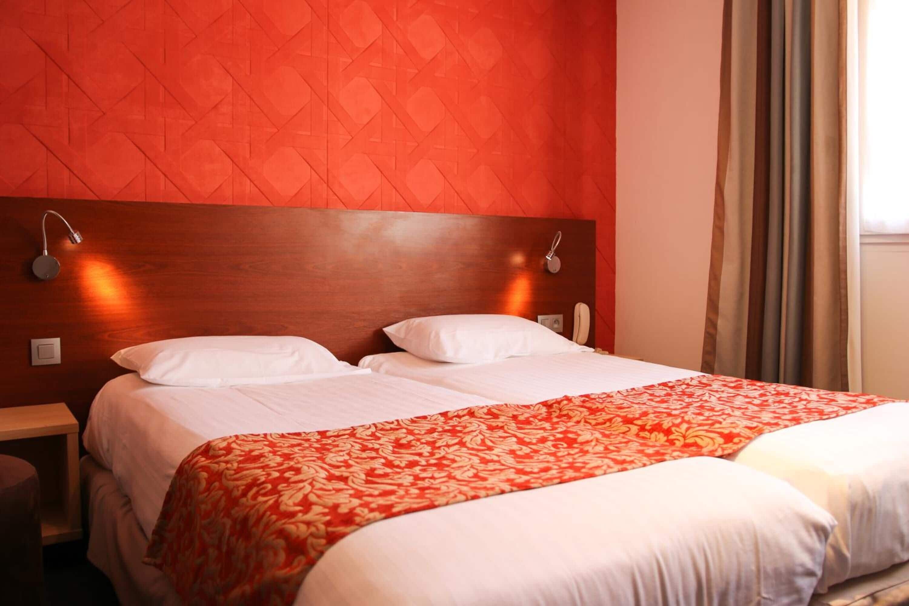 Un lit double avec du linge de lit blanc et une couvre-lit rouge à motifs, une tête de lit en bois et deux lampes de chevet.