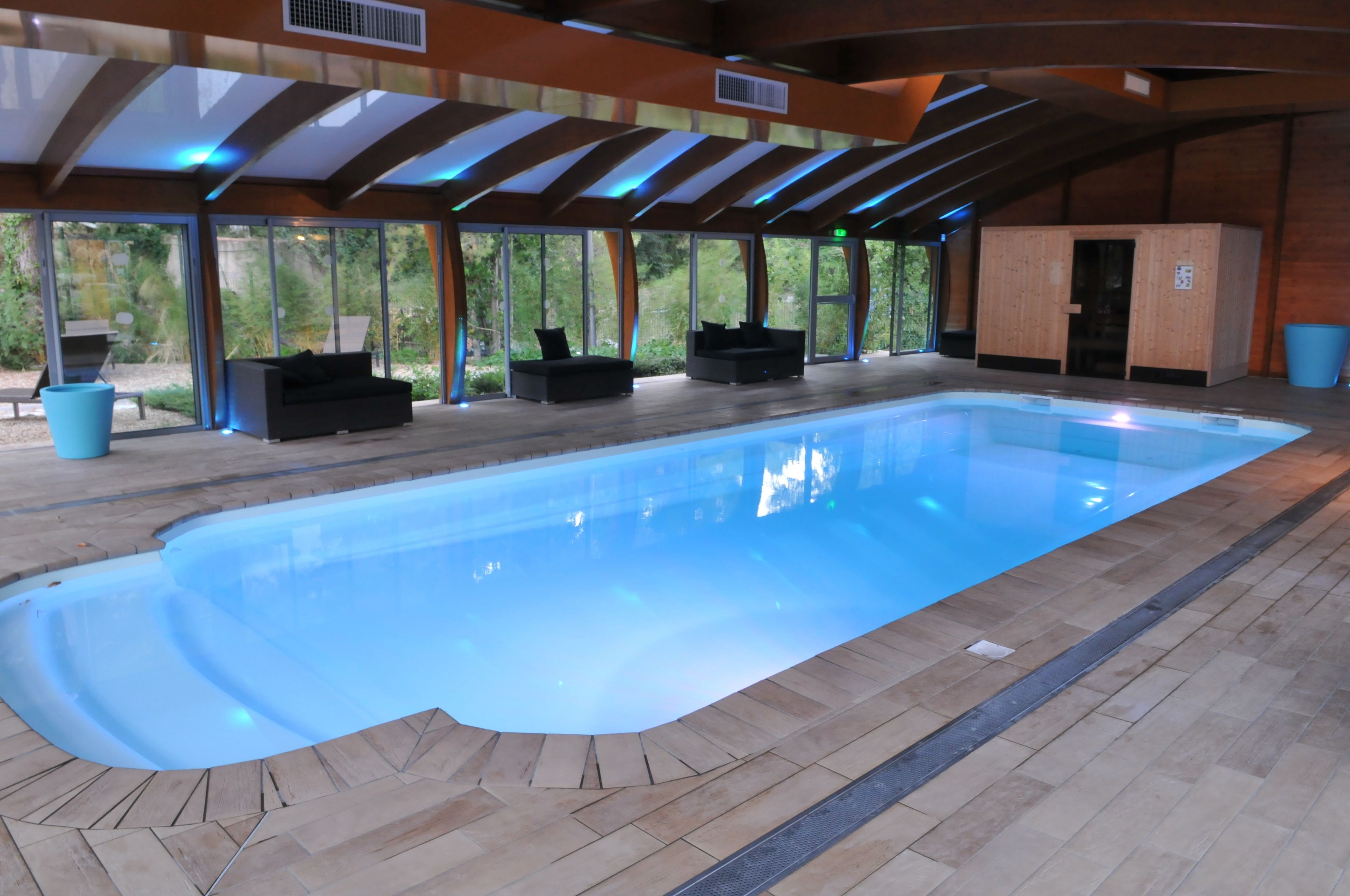 Una piscina interior con una superficie de agua azul cristalina, rodeada de suelo de madera y sillas reclinables negras.