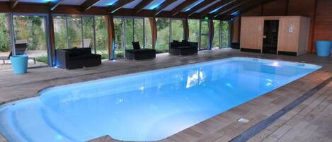 Una piscina interior con una superficie de agua azul cristalina, rodeada de suelo de madera y sillas reclinables negras.