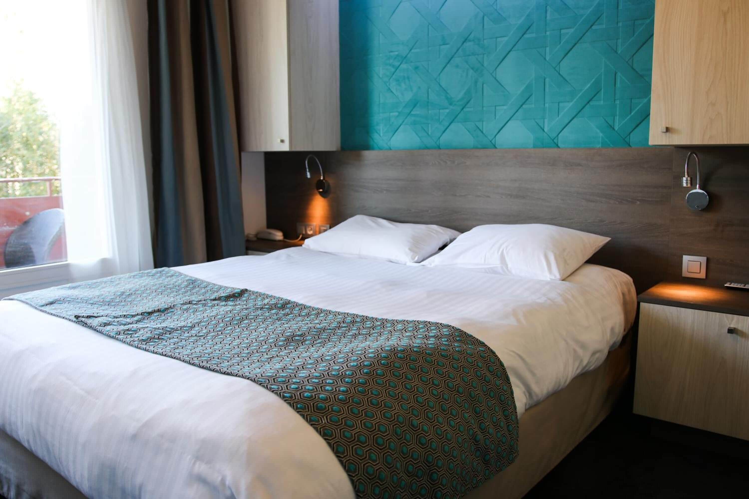 A neatly made bed with a patterned blanket, a bedside table with a lamp, and a wall with a geometric design.