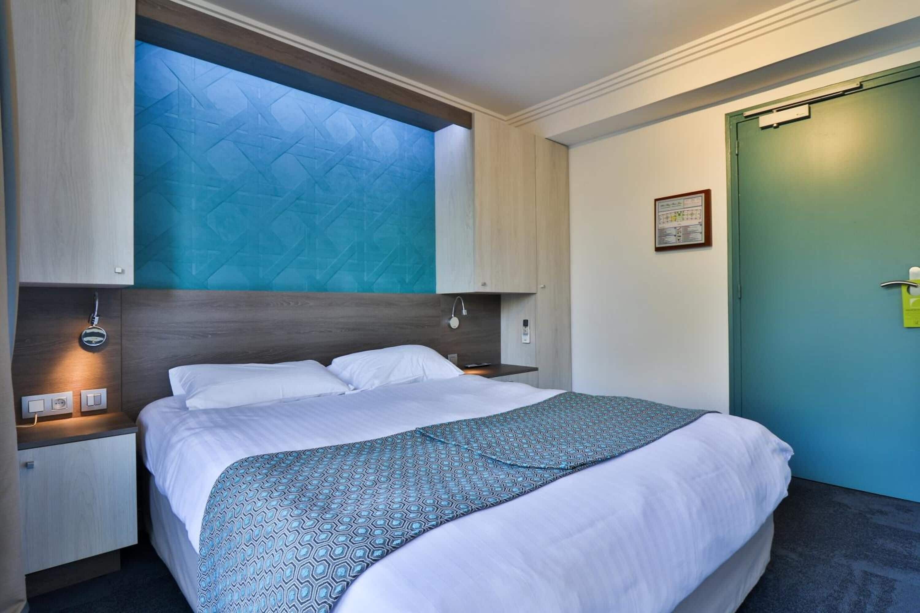 Une chambre d’hôtel moderne avec un grand lit, un puits de lumière intégré, une table de chevet avec une lampe et une porte verte.