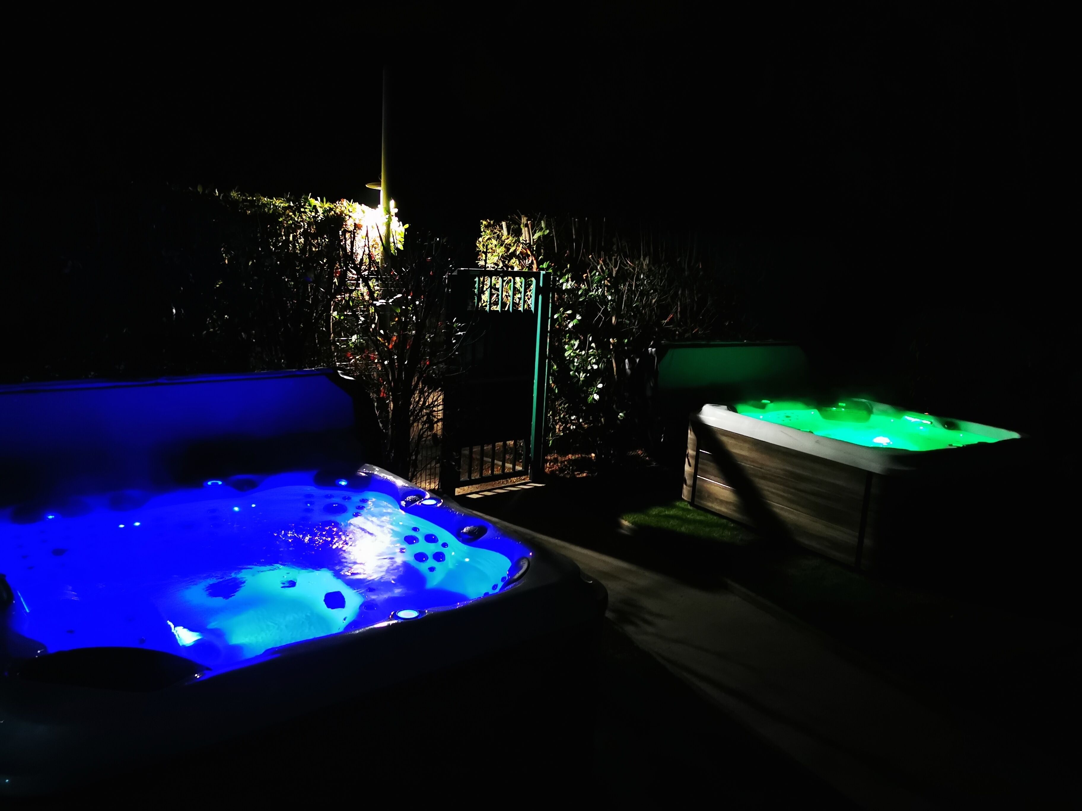A hot tub area with two hot tubs, one with blue lighting and the other with green lighting, surrounded by foliage.