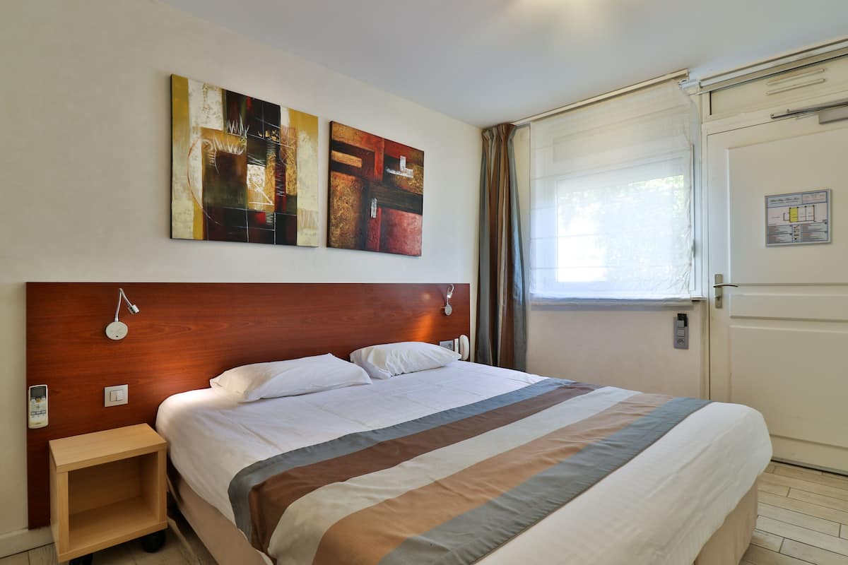 Une chambre d’hôtel avec une tête de lit en bois, un lit avec une literie à rayures, deux tableaux accrochés au mur, une table de chevet avec une lampe et une fenêtre avec des rideaux.