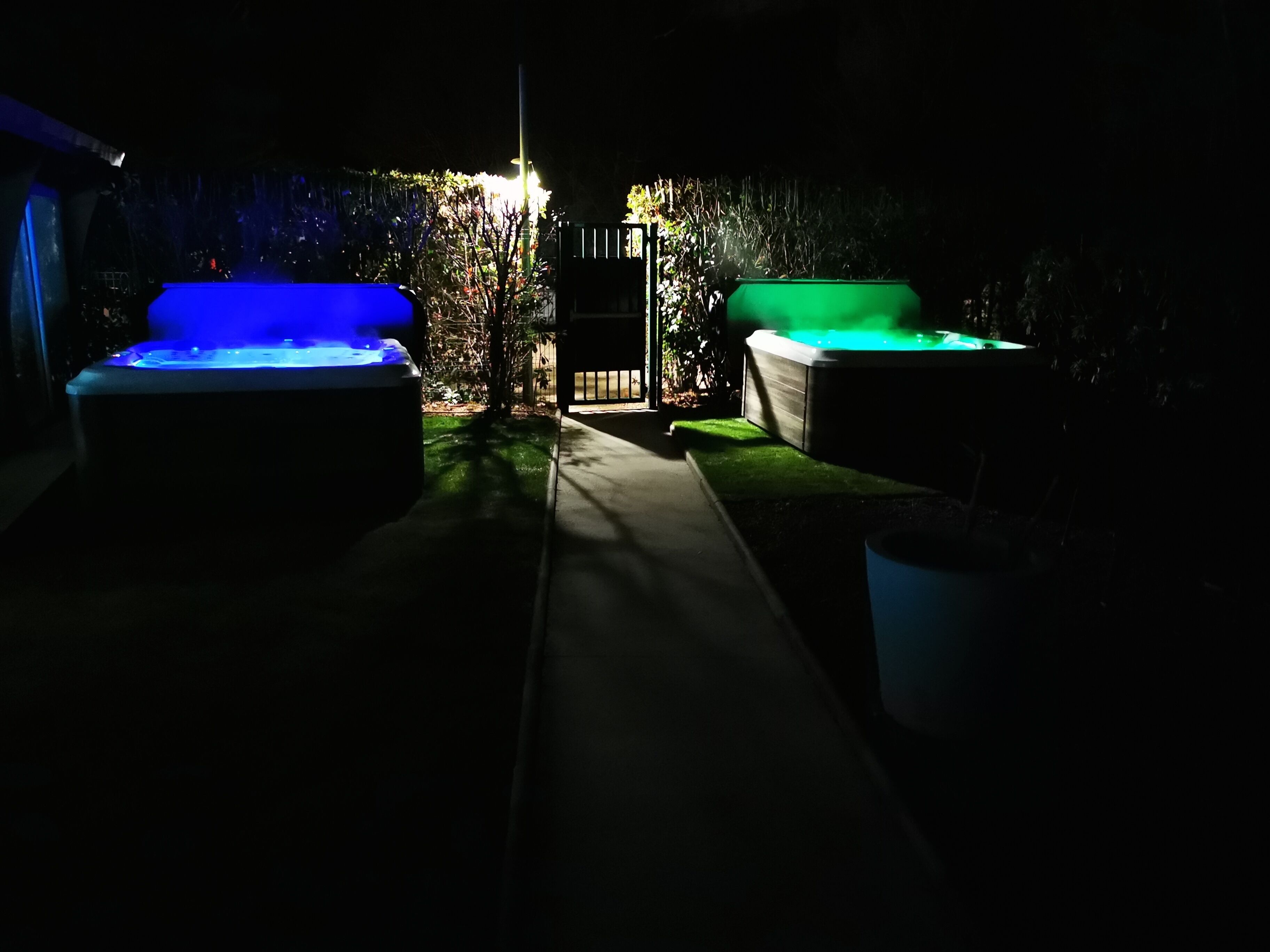 A dimly lit outdoor area with two hot tubs, one with blue lighting and the other with green lighting, separated by a pathway.