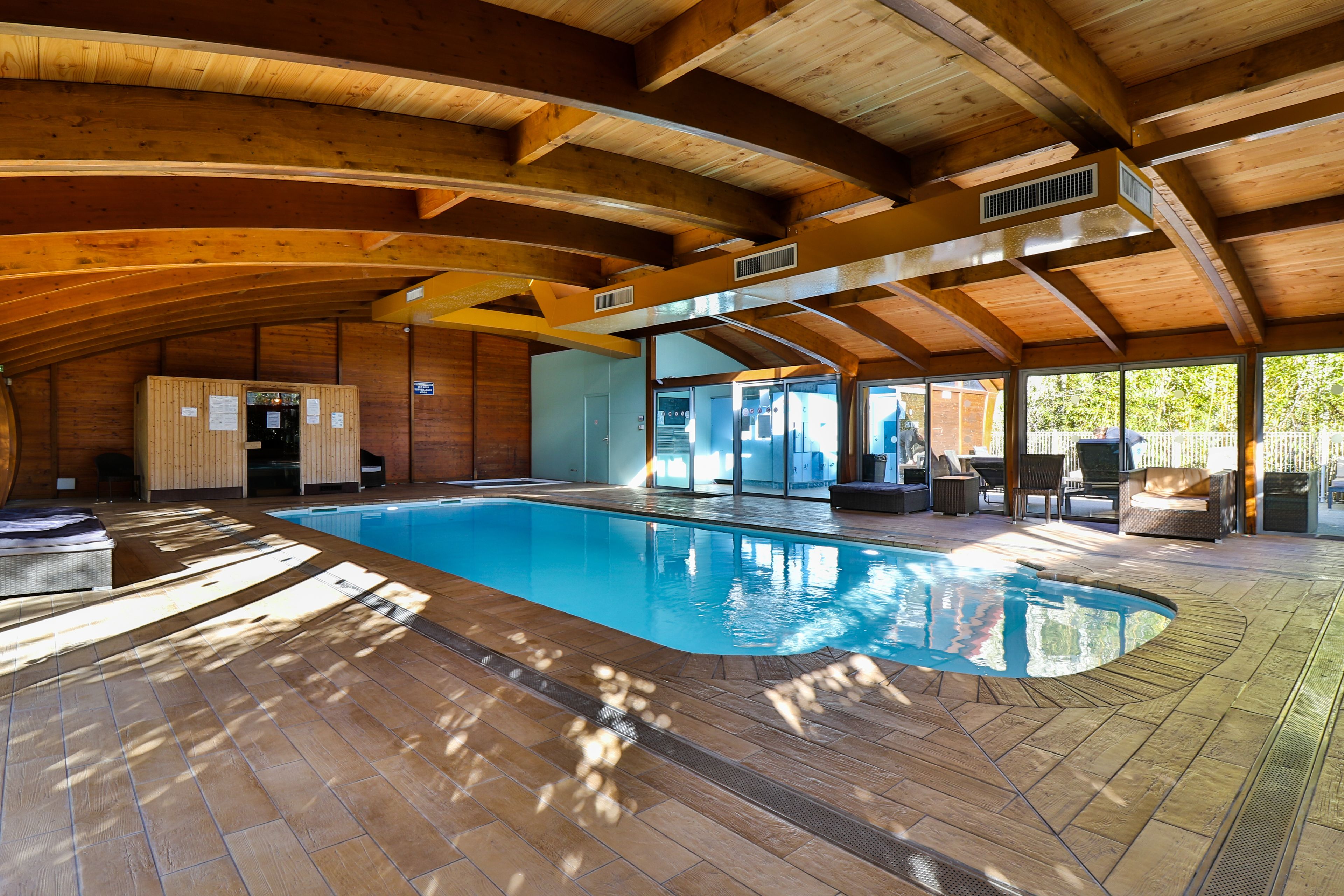 A spacious indoor swimming pool with wooden beams and a tiled floor, surrounded by seating areas and a view of the outdoors.