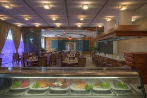 A restaurant with multiple dining tables and chairs, a central chandelier, and a display case with various food items.