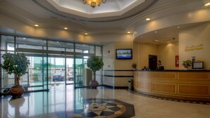 A hotel reception area with a glass door entrance, a reception desk, a television screen, and two potted plants. - Siji Hotel Apartment (Al-Fujairah)