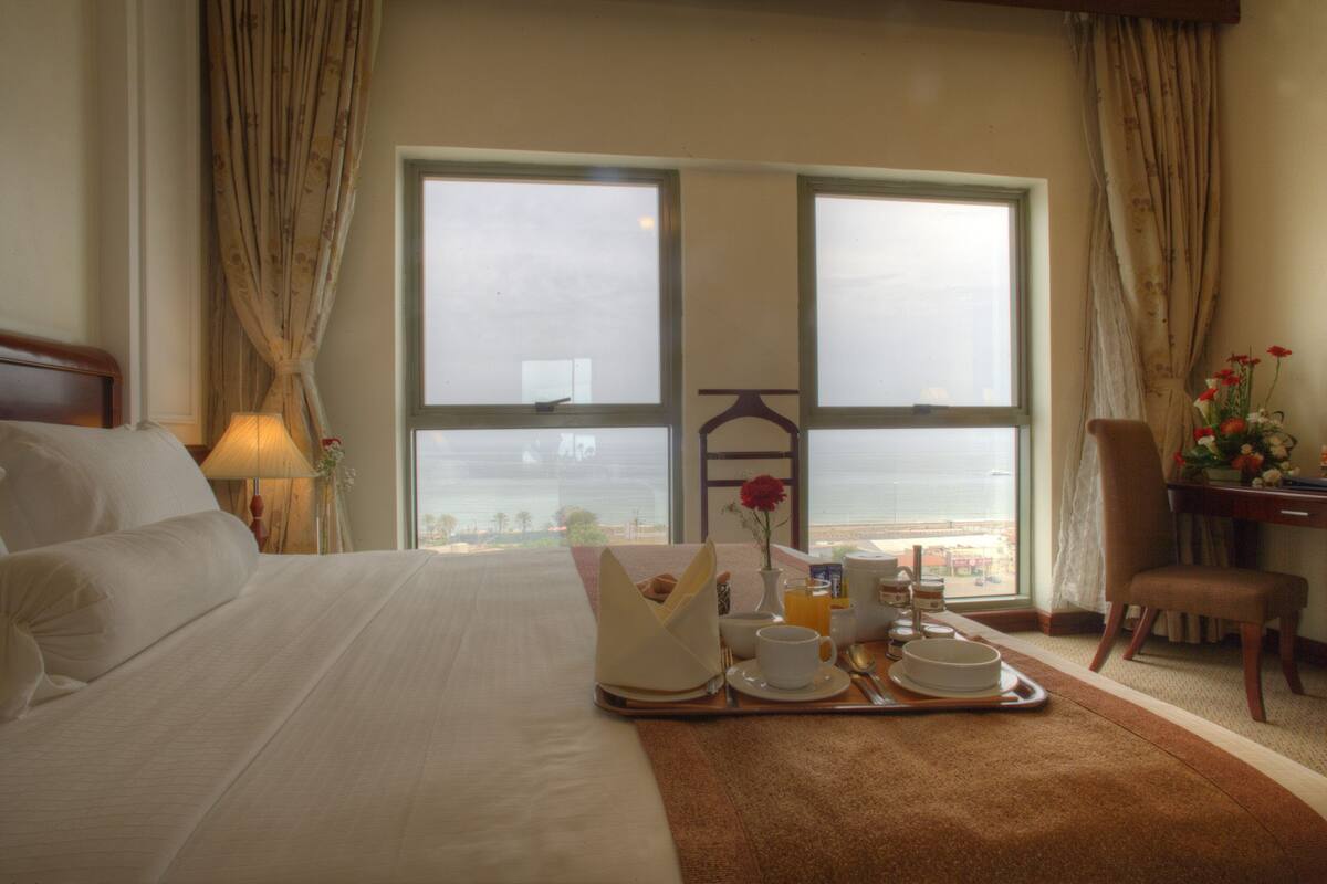 Une chambre d’hôtel avec un grand lit, un plateau de petit-déjeuner, une chaise et une vue sur la mer depuis deux fenêtres.