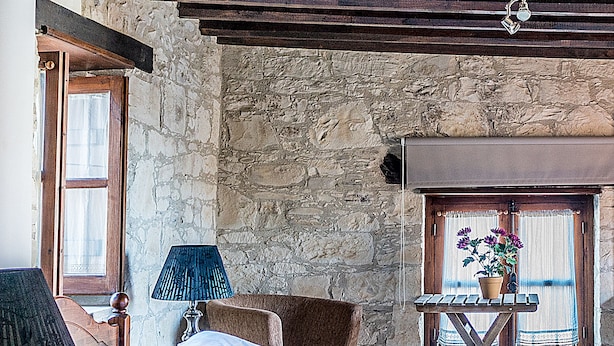 A bedroom with a stone wall, wooden beams, a bed with a floral bedspread, a nightstand with a lamp, and a window with a view.