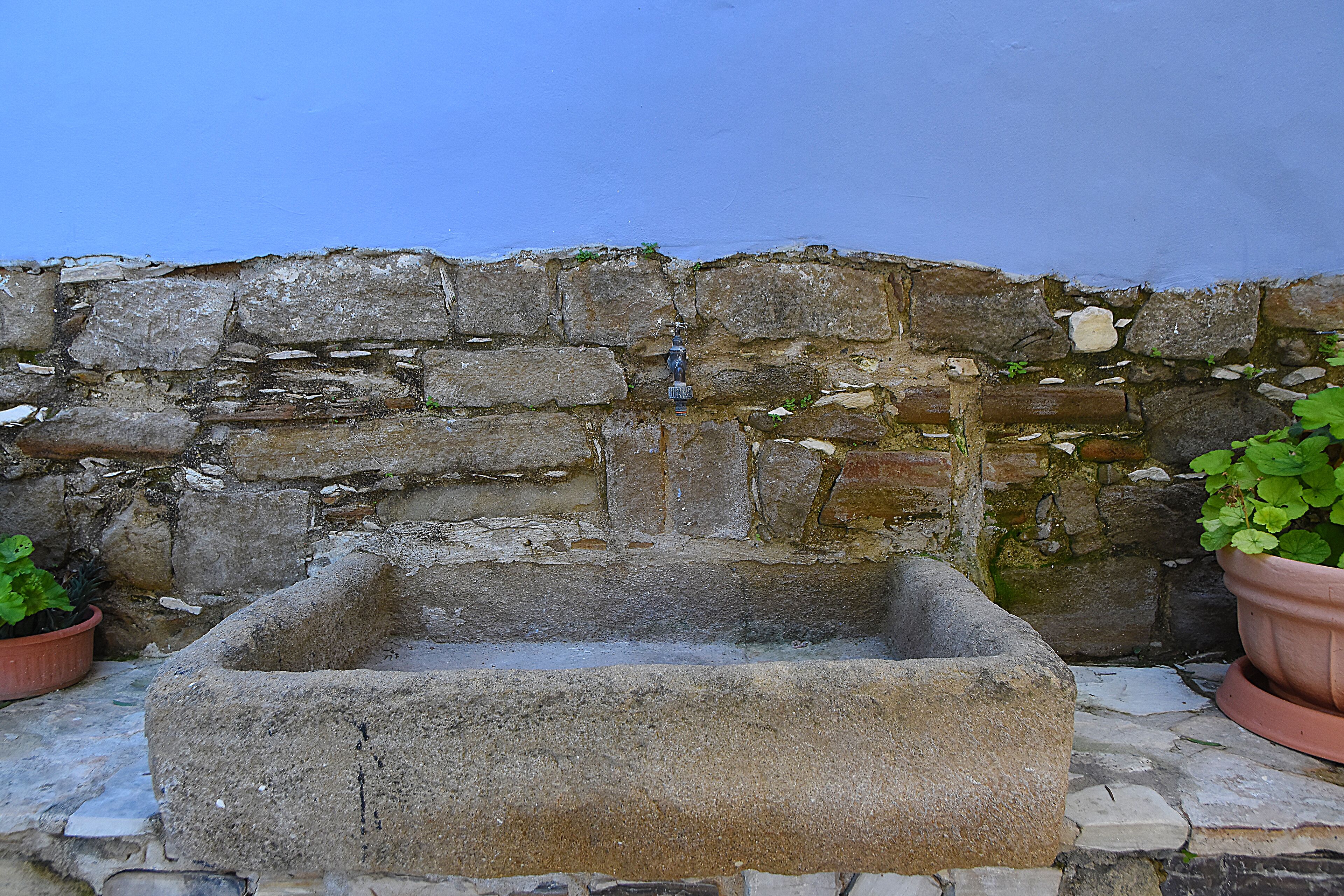 A stone well with a rectangular basin, surrounded by a stone wall with some vegetation growing on it.