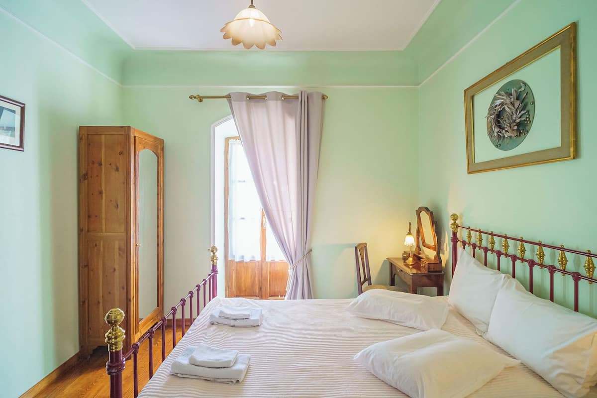 A well-lit bedroom with a large bed, a wooden wardrobe, a window with curtains, and a framed picture on the wall.