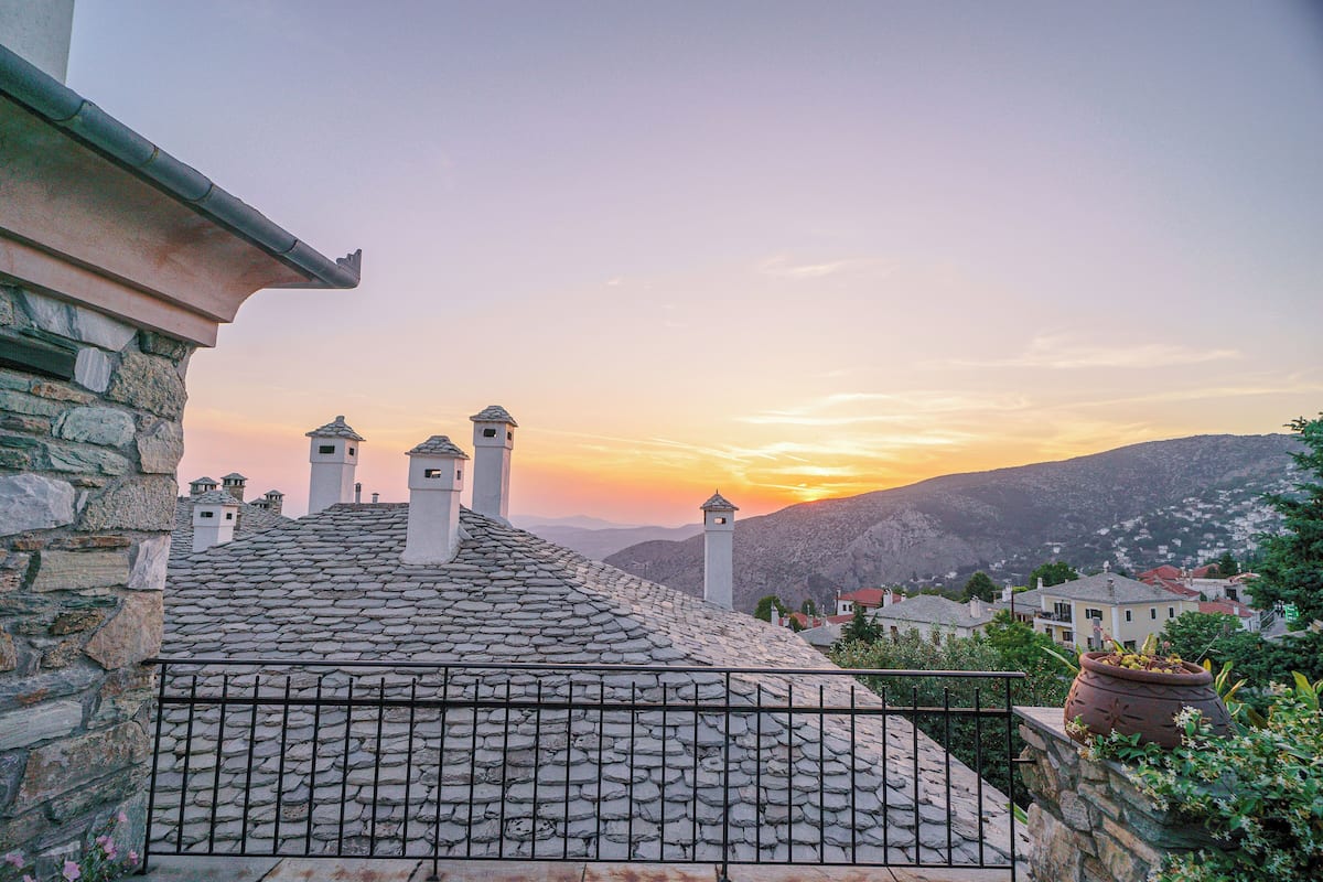 A stone-roofed building with a metal fence, a bell tower, and a view of a sunset over a mountainous landscape.