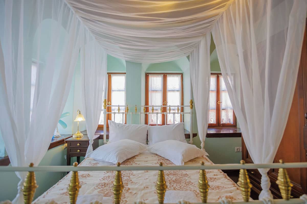 A bed with white linens and a canopy, a bedside table with a lamp, and a window with curtains.