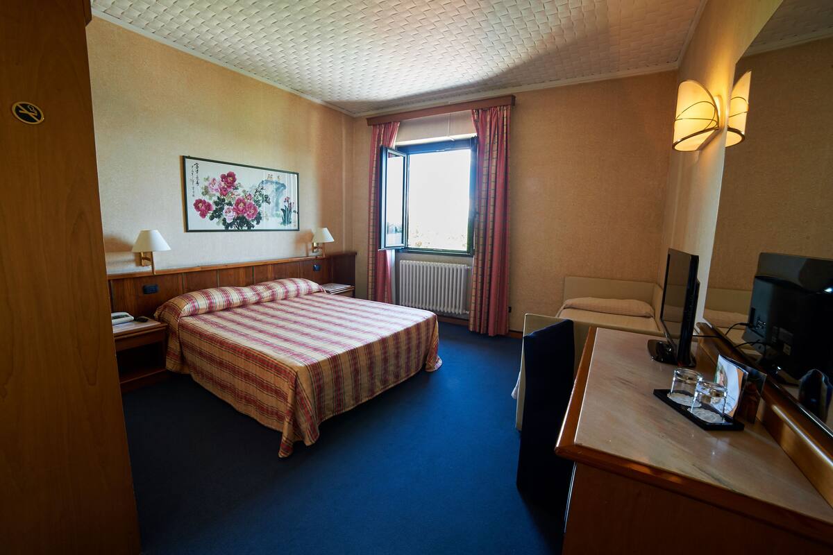 A hotel room with a bed, a desk with a computer, a television, a window with curtains, and a painting on the wall.