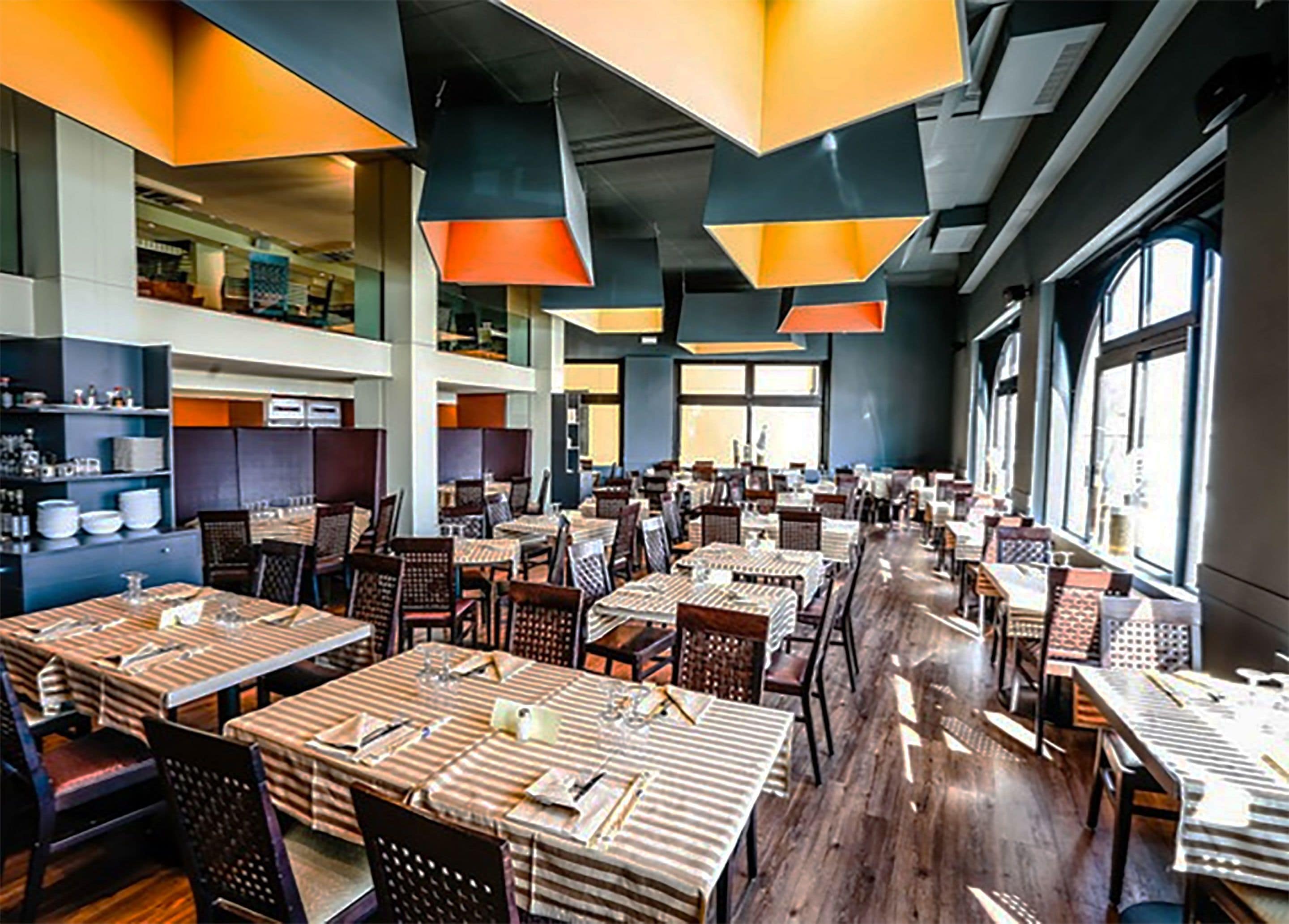Un restaurante amplio con numerosas mesas y sillas, techos altos y grandes ventanales que dejan entrar luz natural.