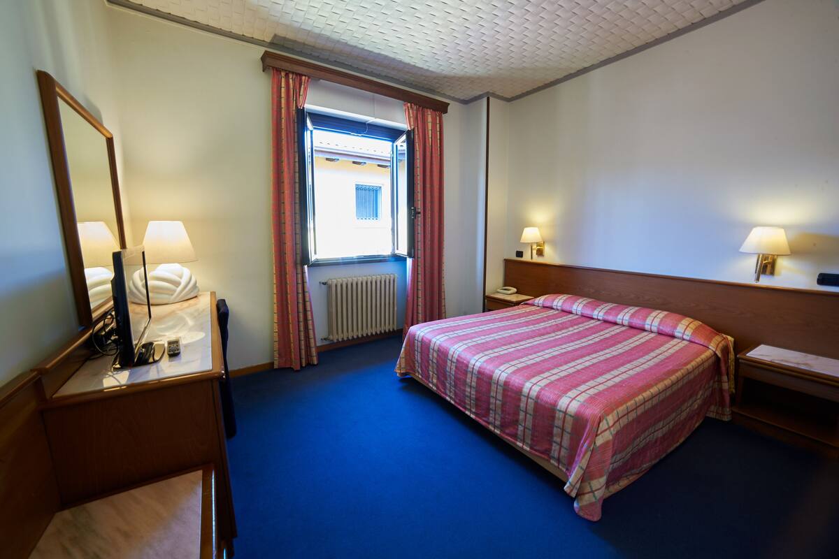 A hotel room with a bed, a desk with a lamp, a chair, a window with curtains, and a radiator.