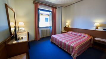 Habitación de hotel con una cama, un escritorio con lámpara, una silla, una ventana con cortinas y un radiador.
