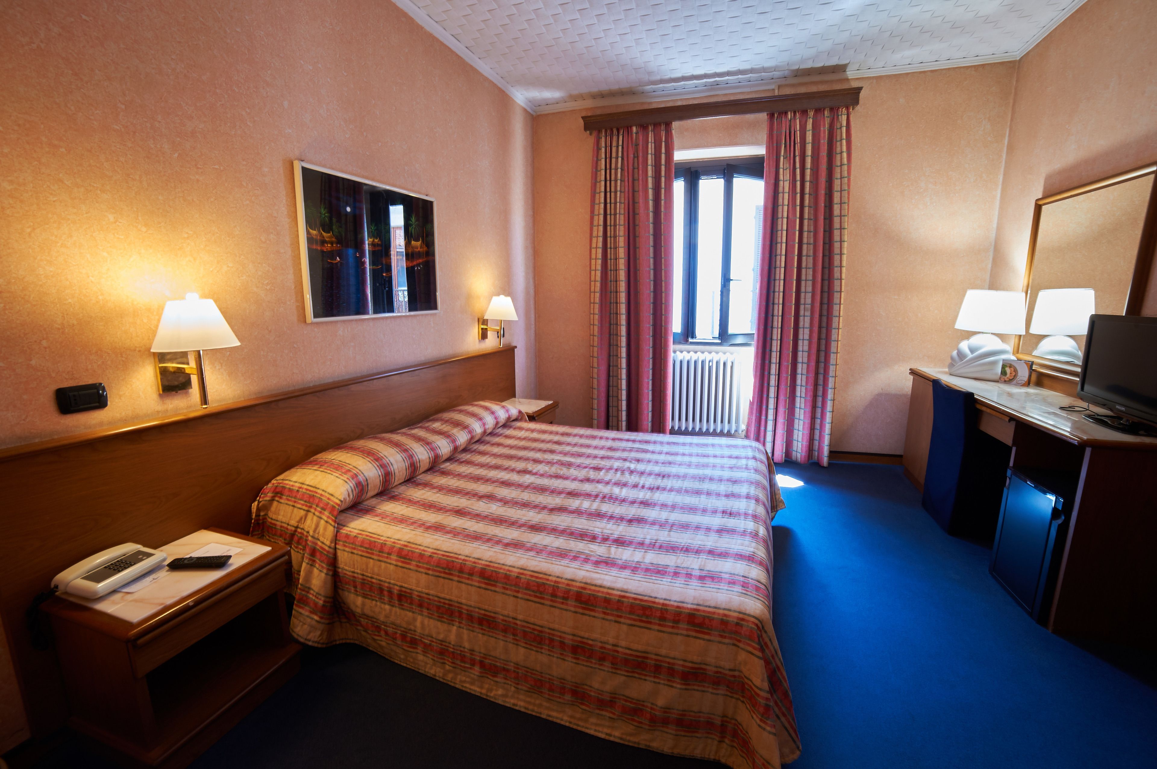 Una habitación de hotel con cama, lámparas de noche, un escritorio con televisor y una ventana con cortinas.